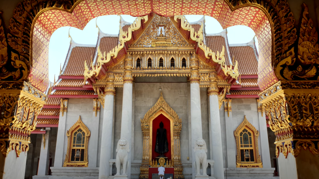 A man in a light colored shirt and shorts praying in front of a white marble Buddhist temple with a red and gold roof.