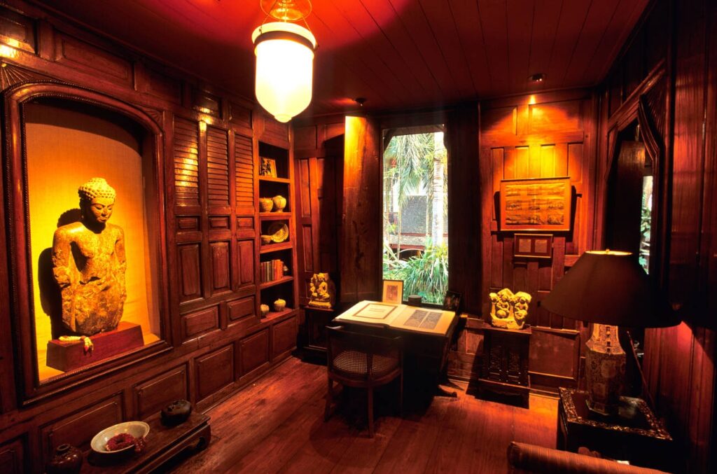 Jim Thompson's office inside his traditional Thai house, featuring dark wood paneling, a Buddha statue, and a writing desk.